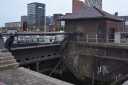 a large hydraulic-operated canal lock with apartment buildings in the background.