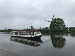 an enclosed canal boat with large windows motoring done a canal, with dutch windmills in the background