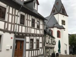 a street with half-timbered houses on the left, leading to a church with large clock in the steeple tower.