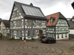 A historic half-timbered house with solar panel on the roof, sitting at the edge of a cobblestone courtyard